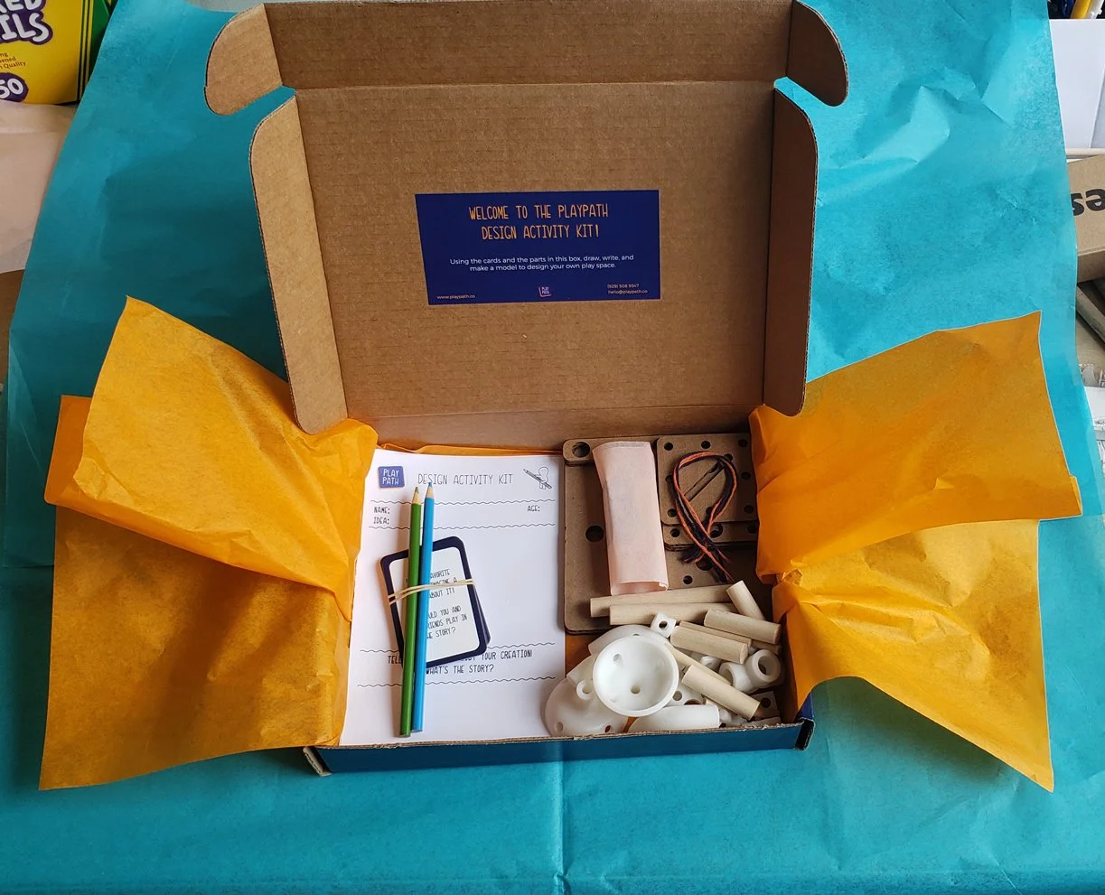 Open cardboard box with yellow tissue paper, containing a design activity kit with pencils, wooden pieces, a small motor, and instructions on a sheet of paper, on a blue surface.