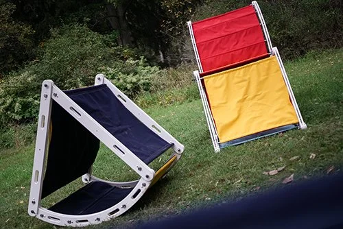 Two foldable lawn chairs, one black with white frame on the ground and another red and yellow chair leaning on grass, in a backyard setting.