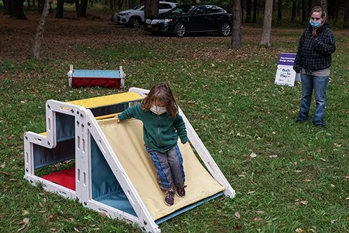 Child sliding down an outdoor playground slide with a woman standing nearby in a park, both wearing masks.