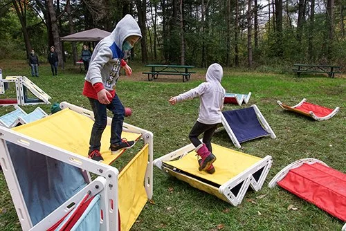 Two children playing on and around colorful wooden ramps in a park with trees, grass, and picnic tables.