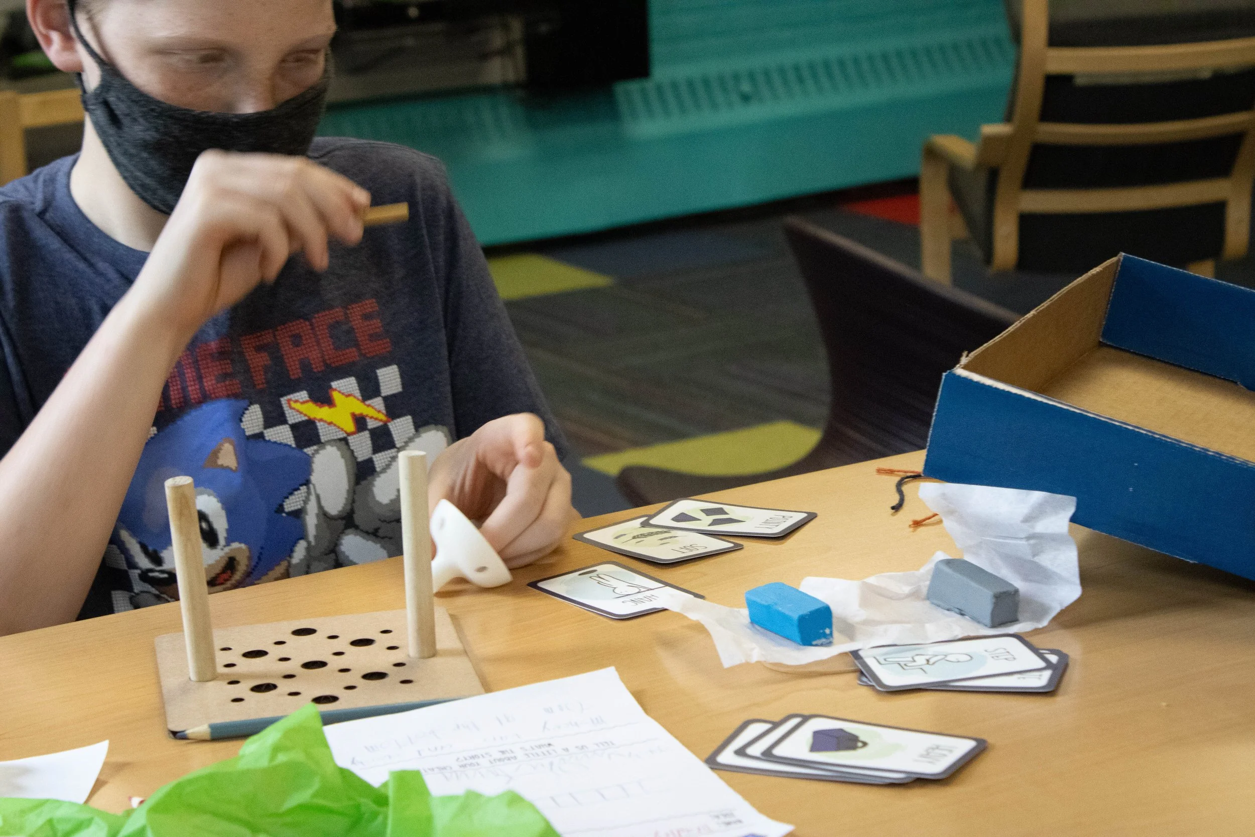A young boy with a face mask playing a tabletop game with cards and game pieces on a table in a classroom or activity room.