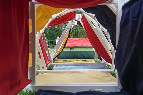 View inside a colorful children's play tent with red, yellow, black, and blue fabric panels and a padded floor, outdoor green scenery seen through the open entrance.