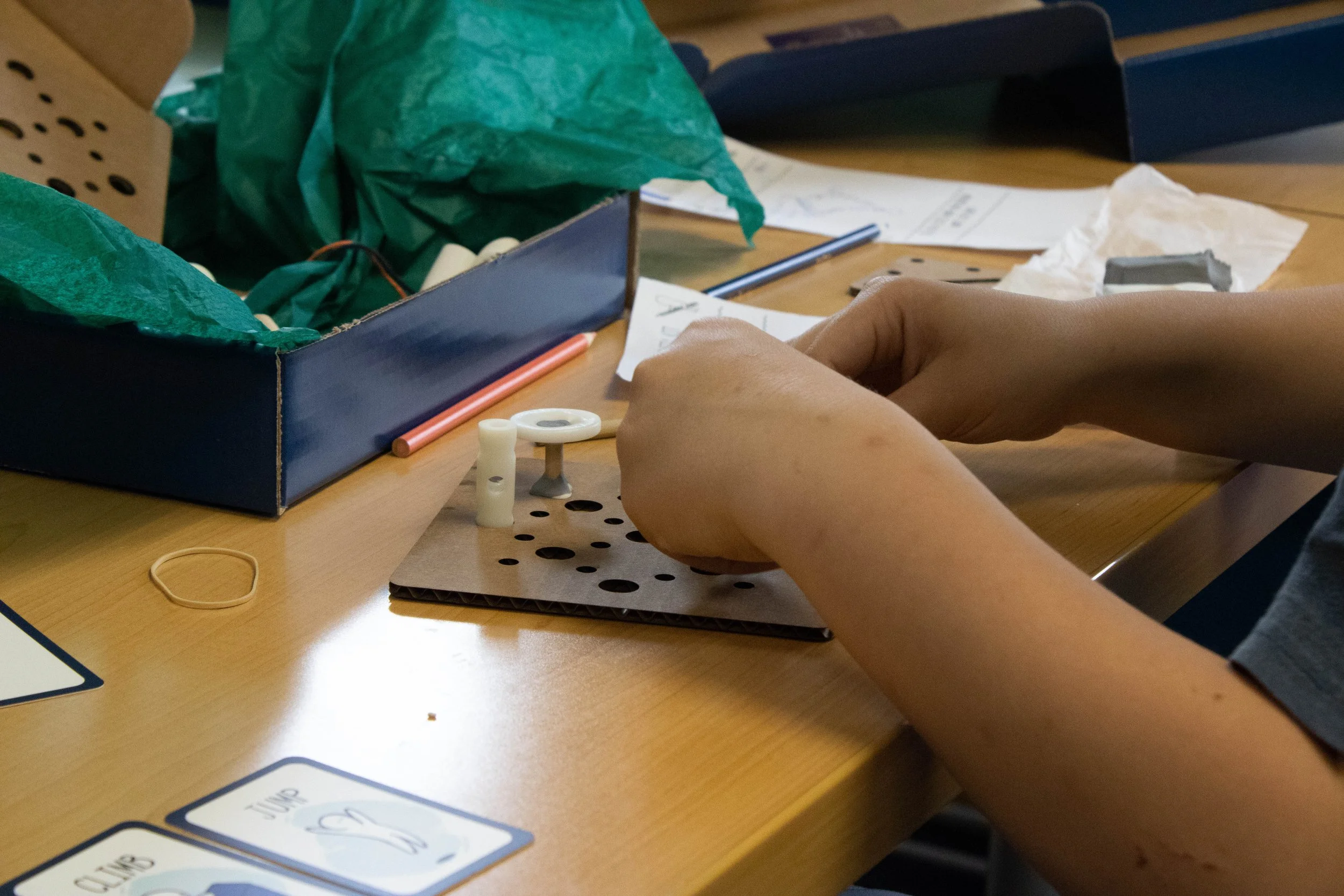 Person's hands working with medical or scientific supplies on a wooden table, with papers, a pen, and small plastic parts nearby.
