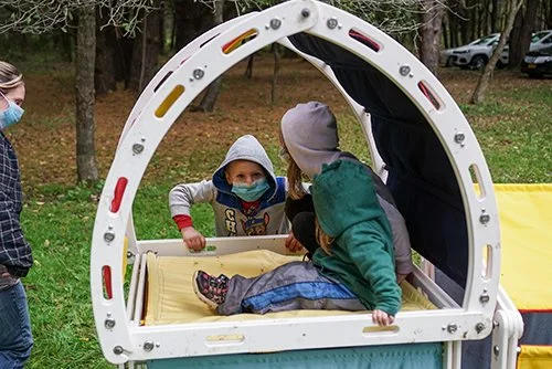 Children at a playground, with one child lying on a yellow padded surface inside a white arch-shaped play tunnel, while two other children stand nearby, wearing masks and winter clothing, in a wooded outdoor area.