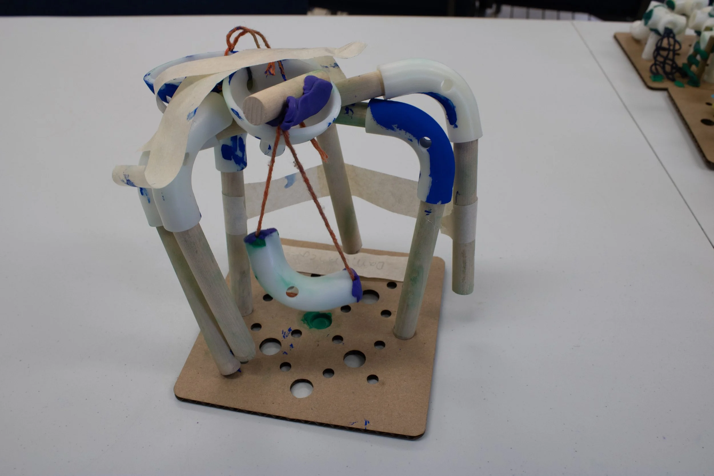 A small wooden and plastic mechanical structure with pipes, strings, and painted components on a white surface.