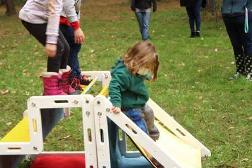 Child climbing on a yellow and white playground slide in a grassy area with adults in the background.