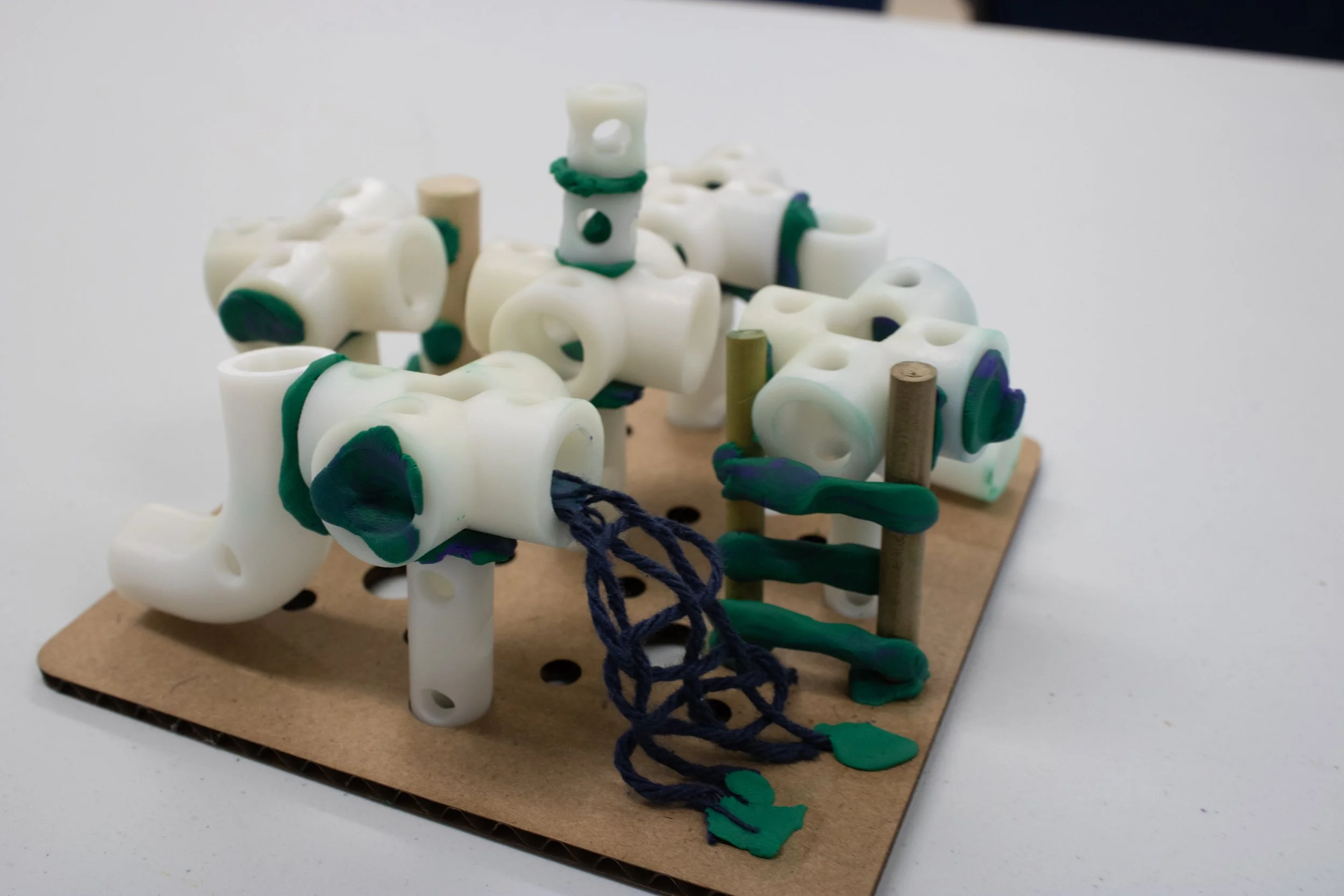 A small model or sculpture of a structure made from white plastic parts connected with green and purple rubber bands, mounted on a piece of cardboard.