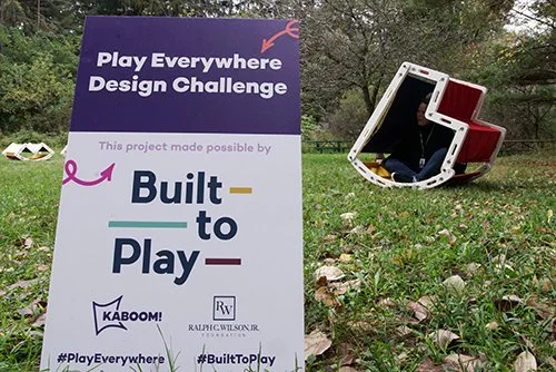 Outdoor scene with a sign promoting the Play Everywhere Design Challenge and a large overturned red and white seat on the grass.