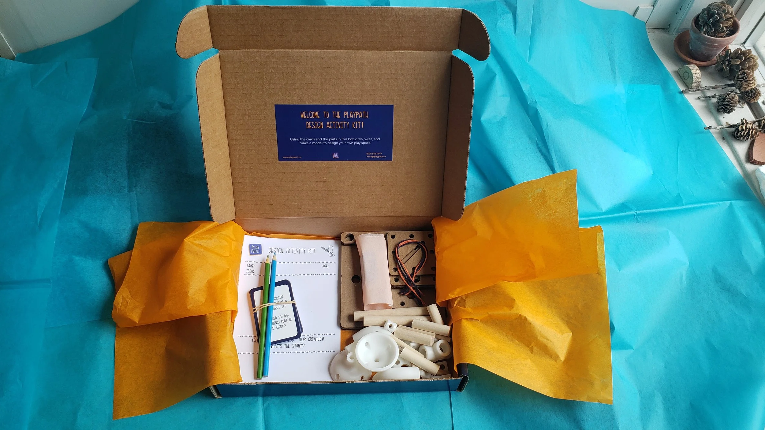 Unopened cardboard box labeled 'Playpath Design Activity Kit' on a blue table with yellow tissue paper, craft materials, and pinecones nearby.