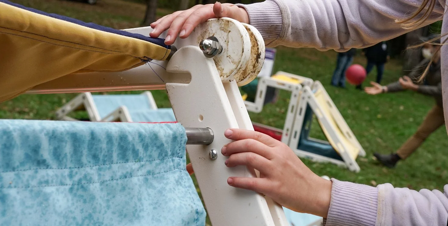Child and adult working together to assemble a piece of outdoor furniture, with various window frames and children playing in the background.