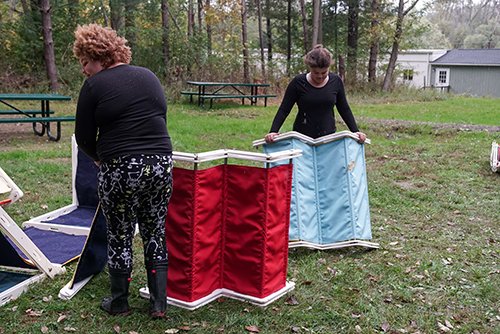Two women setting up portable foldable privacy screens outdoors in a grassy area with trees and picnic tables.