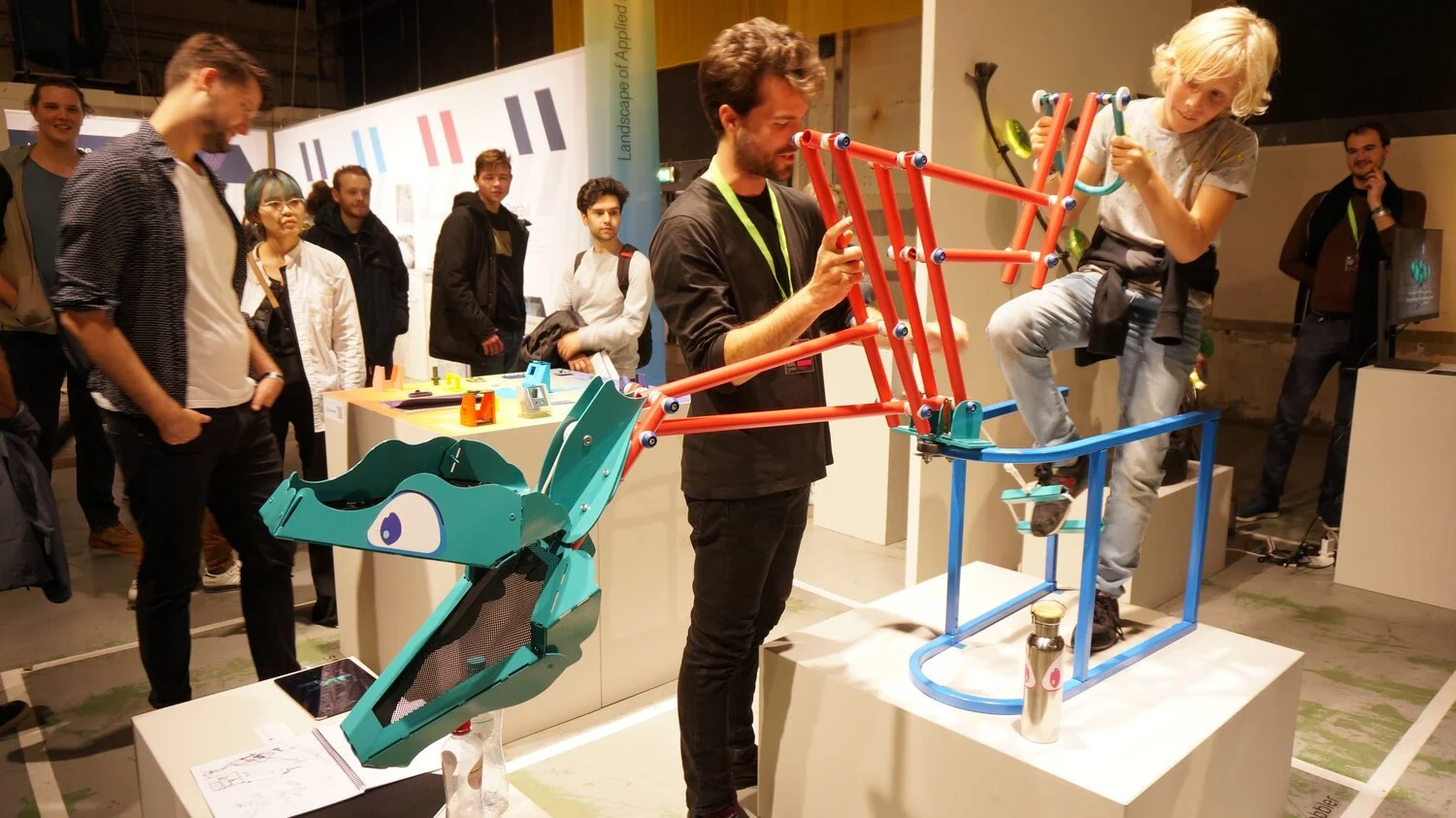 A child is balancing on a colorful, robotic playground structure at a science or innovation exhibit with several onlookers observing.