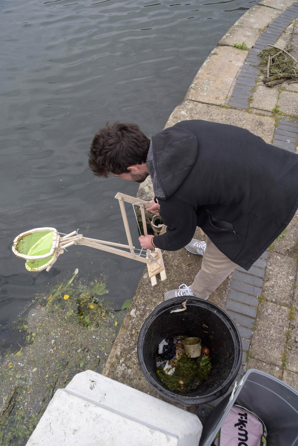 Man collecting water samples using a device by a riverbank with a bucket and debris.