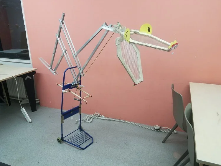 A robot designed to look like a dinosaur, made with various scraps and mechanical parts, standing in a room with pink walls, next to desks and chairs.