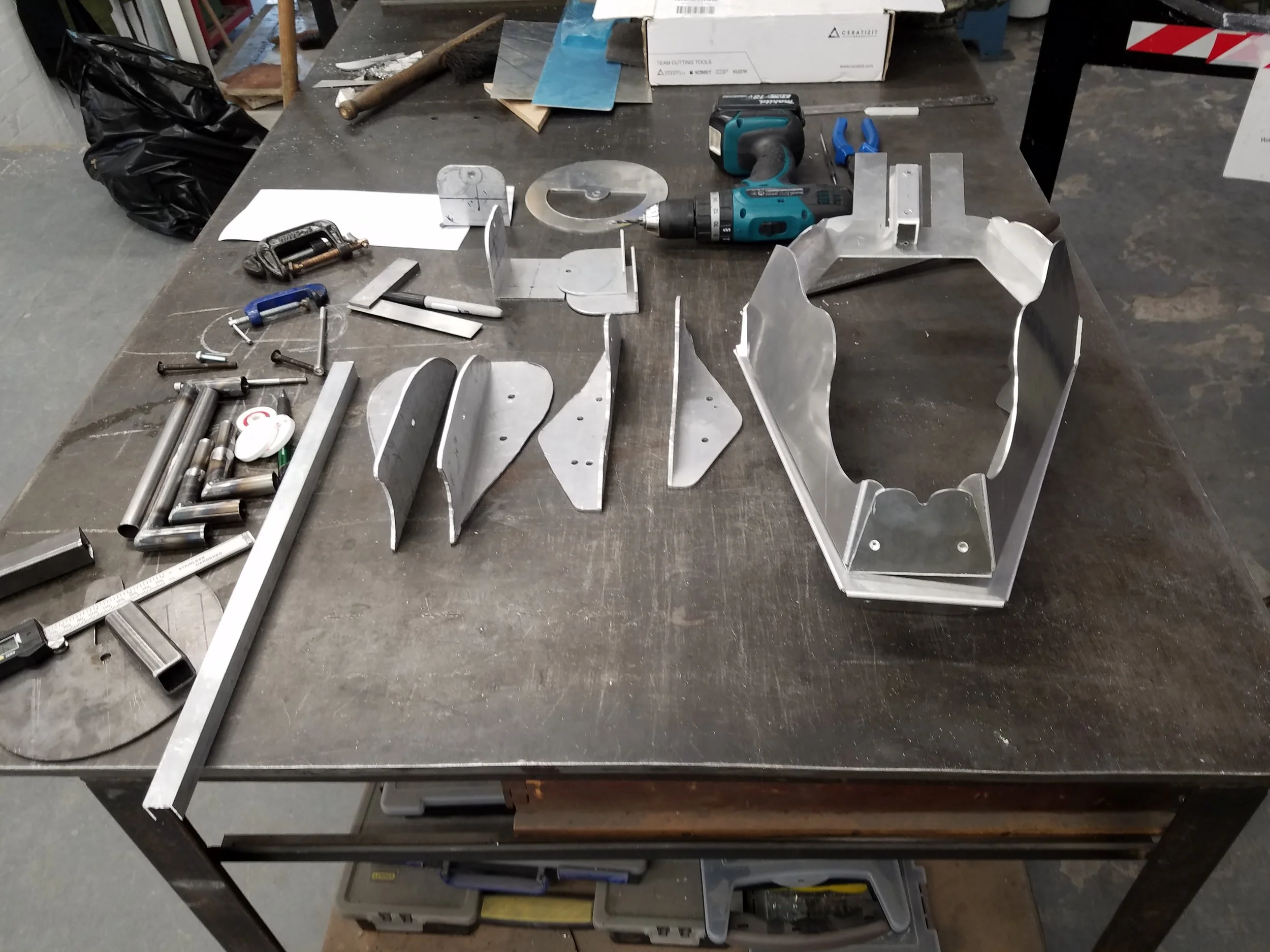 A cluttered workshop table with metal parts, tools, a cordless drill, and miscellaneous hardware. In the background, there are shelves and a black trash bag.