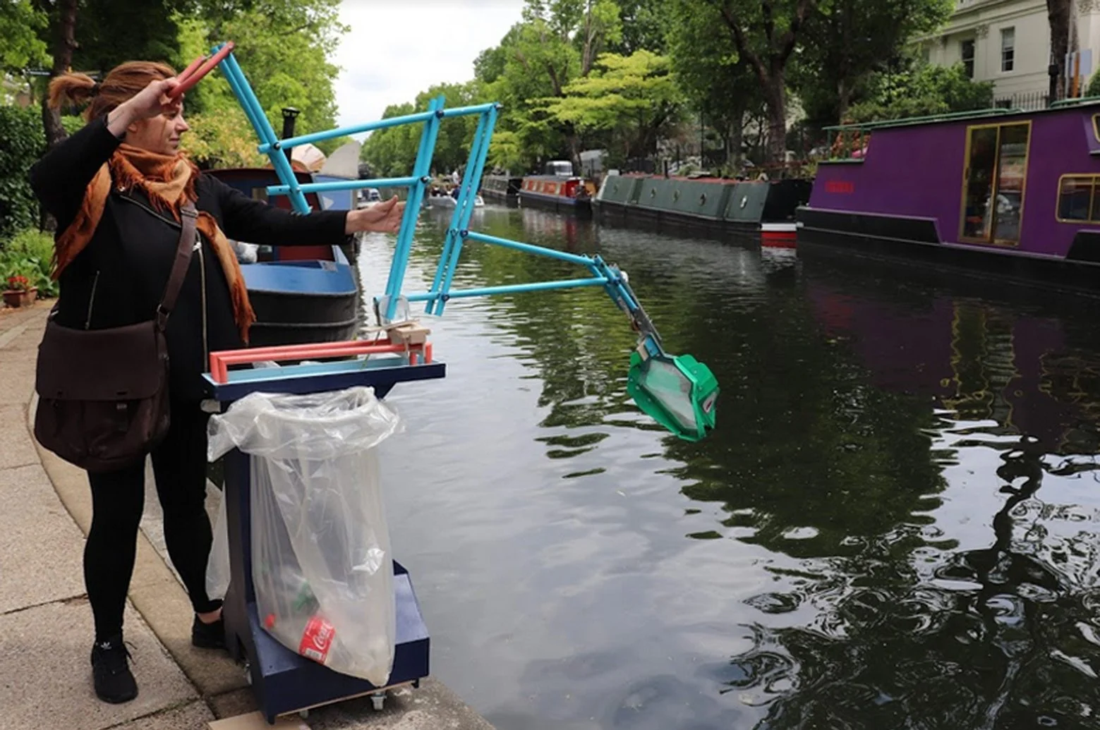 A woman on a sidewalk near a canal, holding a small blue and orange boat with her right hand, and appearing to throw or retrieve something from the water. There are colorful boats docked along the canal in the background, with trees and buildings lin