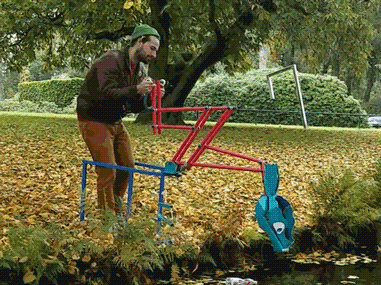 A man is looking at his phone while standing next to an upside-down red and blue prop guitar on a playground with fallen leaves, trees, and bushes in the background.