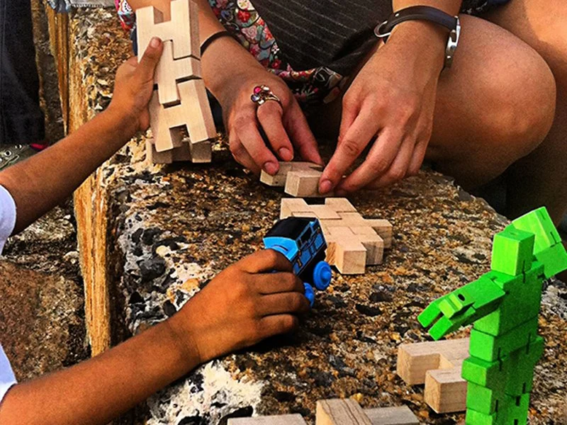 Two children and an adult playing with wooden puzzle pieces and LEGO toys on a stone surface.