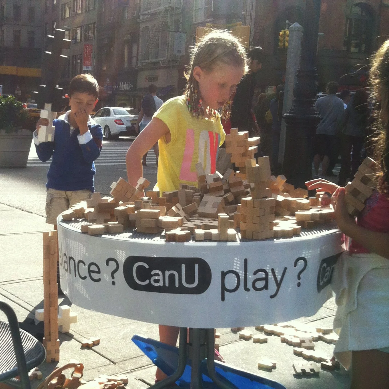 Children playing with wooden blocks at a street table, with a sign asking, "Can U play?" and a busy city street in the background.