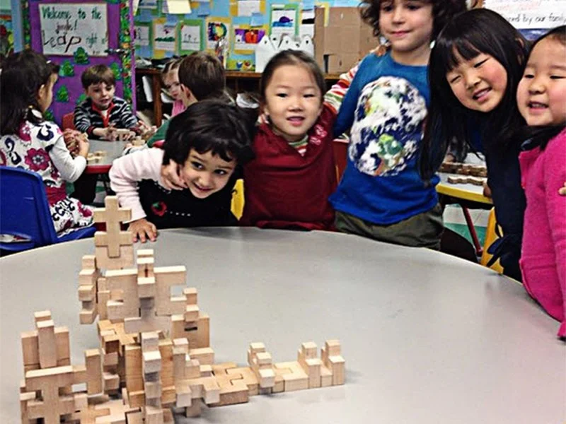 Group of children gathered around a table watching a tower of wooden blocks with a small cross on top, inside a classroom with colorful art displays in the background.