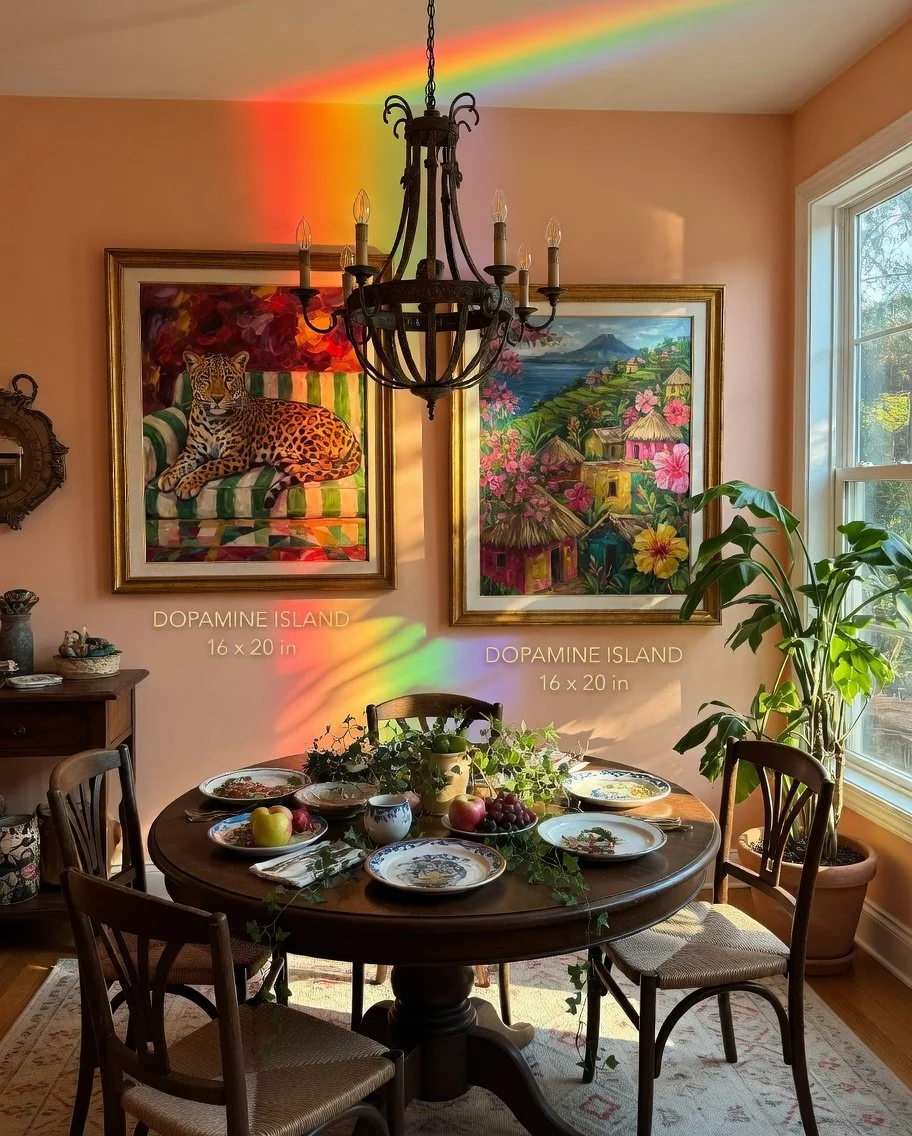 Sunlight passing through a window creates a rainbow on the wall. The room has a round wooden dining table with china plates, apples, grapes, and a potted plant, surrounded by four wooden chairs. Two colorful paintings hang on the peach-colored wall behind the table, one featuring a leopard lounging on a striped chair and the other depicting a hillside village with flowers. A black chandelier hangs above the table, and a large potted plant is near the window on the right.