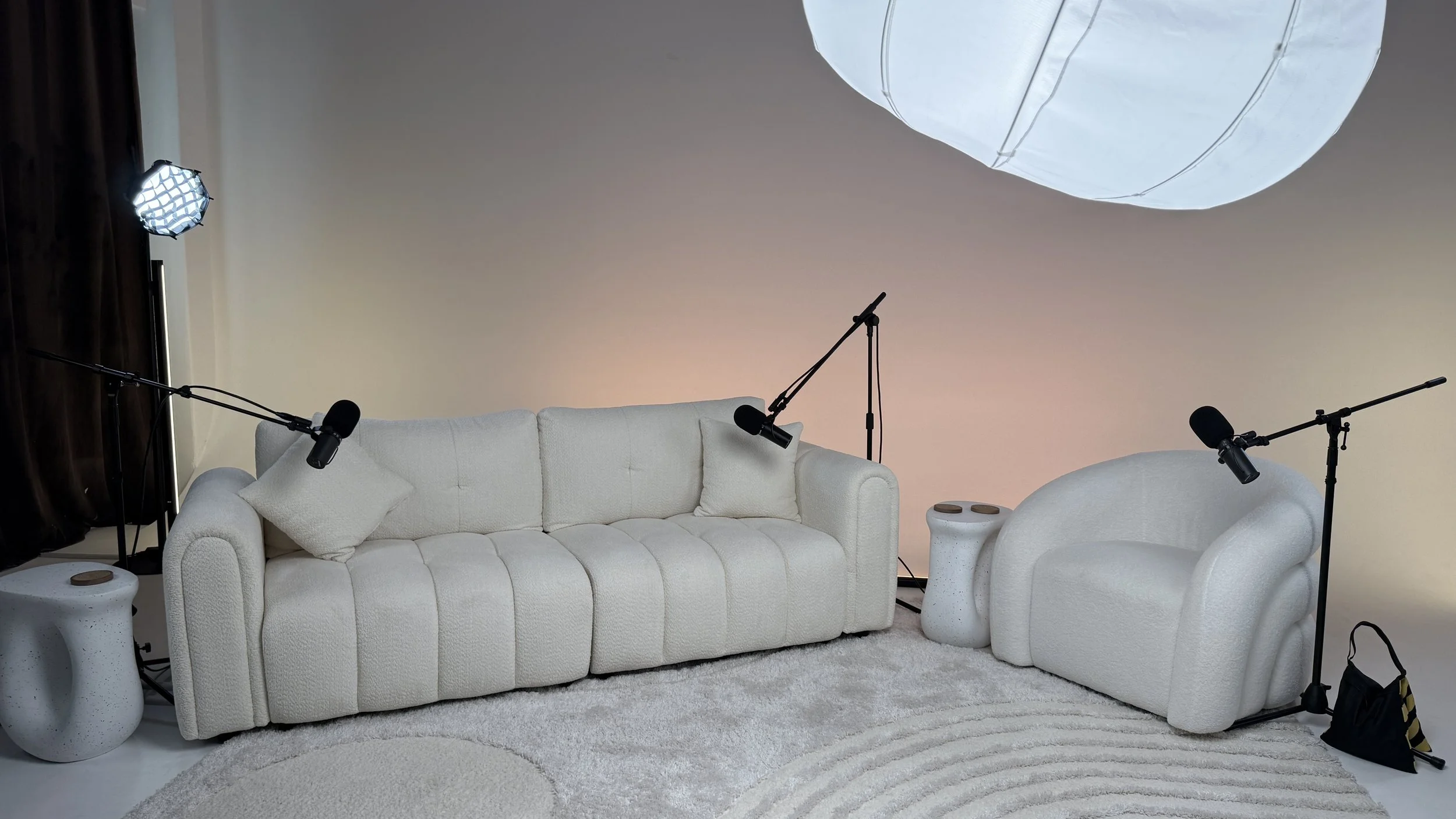 Three-person podcast set with white sofa, lounge chair, side tables, and boom microphones