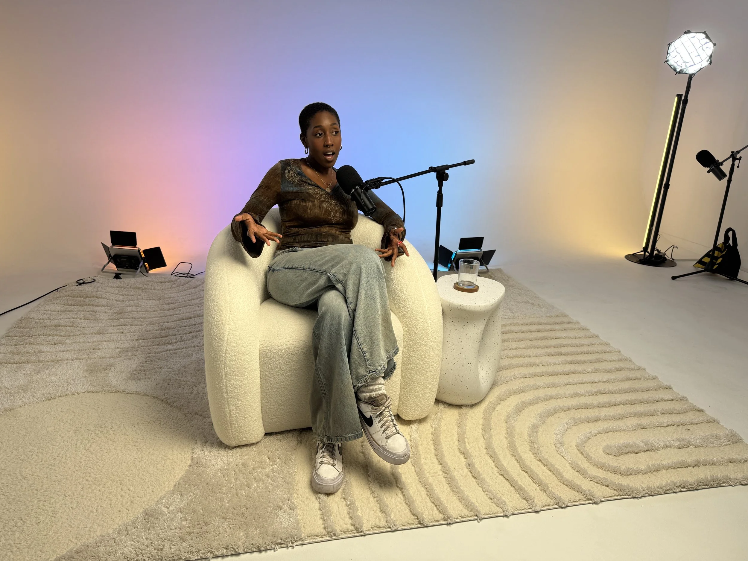 Podcast guest speaking into a microphone on a white lounge chair with colorful lighting behind her