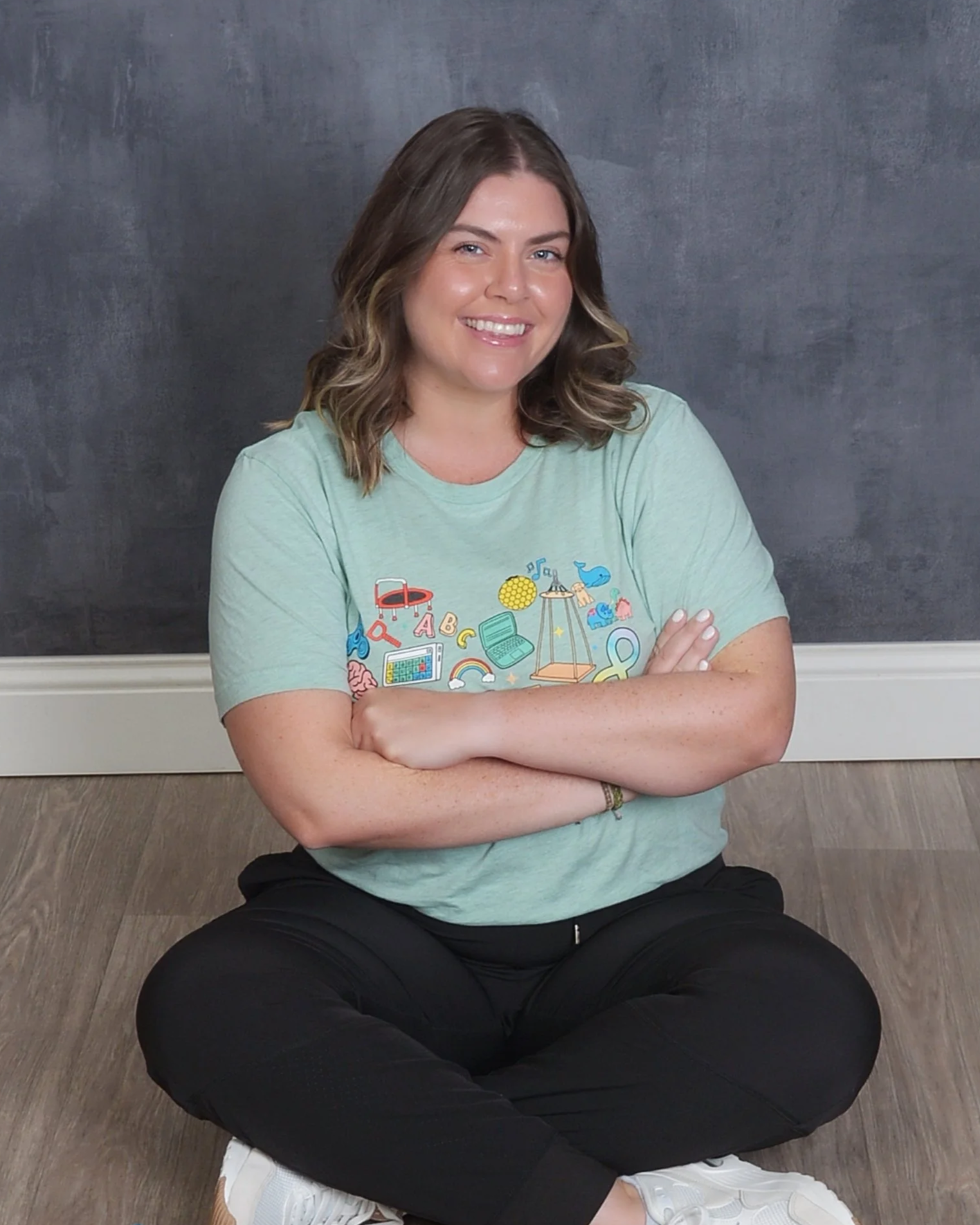 A speech therapist woman with shoulder-length wavy brown hair sitting cross-legged on a wooden floor in front of a dark gray wall, smiling and wearing a light green graphic T-shirt and black pants.