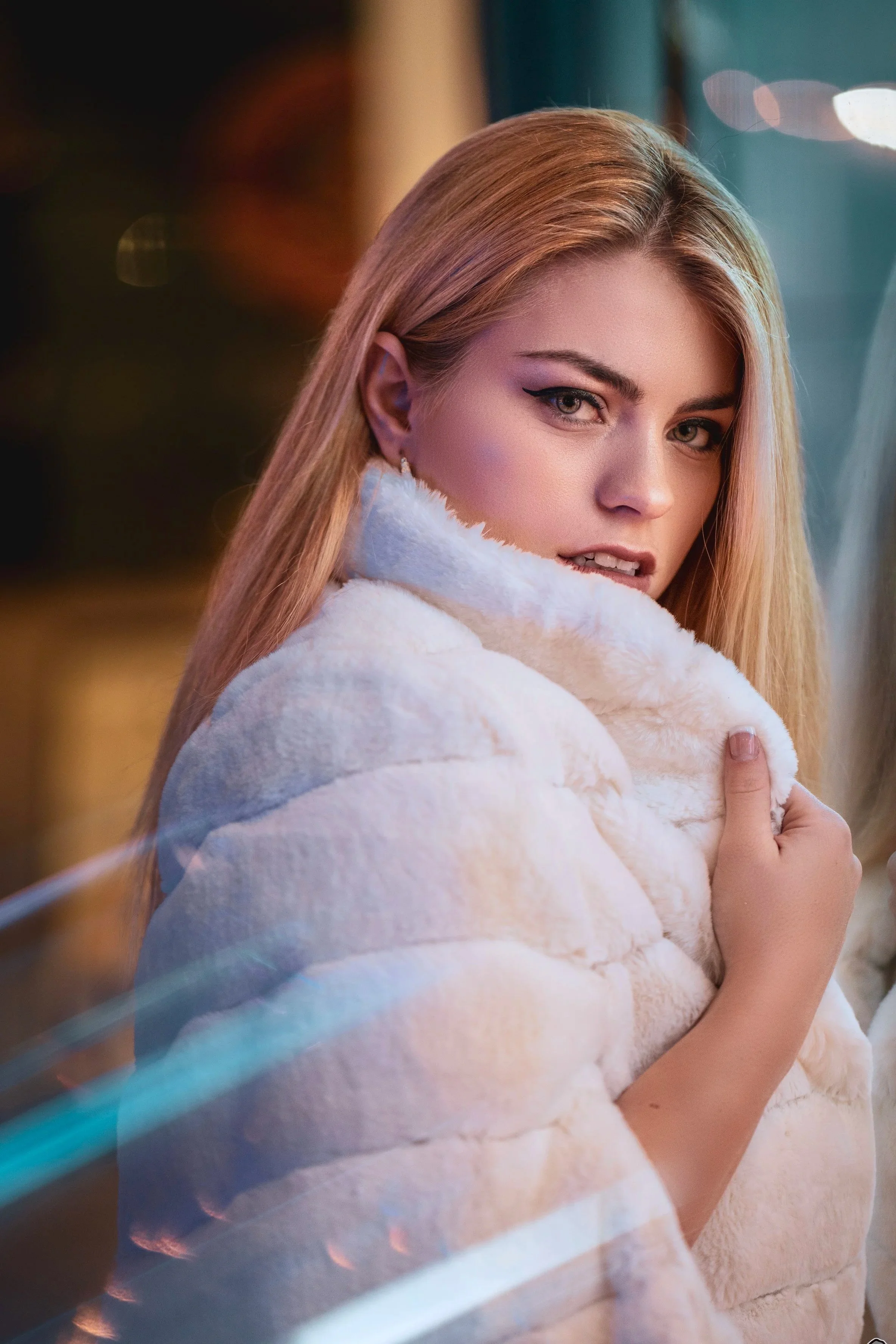 A woman with blonde hair and grey eyes wearing black eyeliner, a white fur coat, and earrings looking over her shoulder indoors with lights in the background