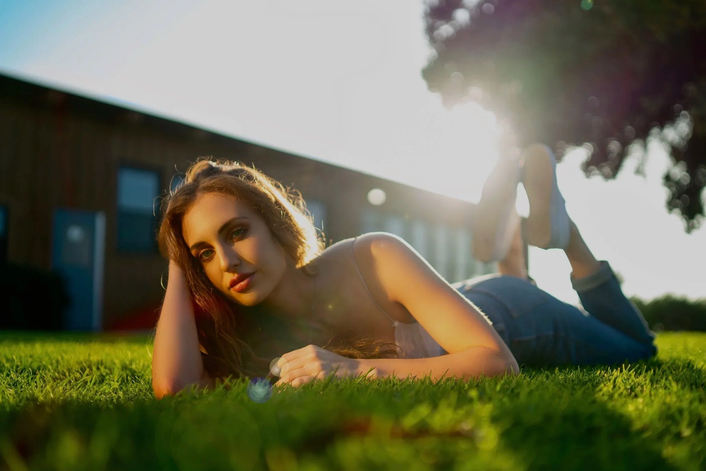 A young woman with red hair lying on her stomach on a grassy lawn, looking at the camera with her head resting on her arm. The sunlight is bright behind her, creating a glow and lens flare effects.