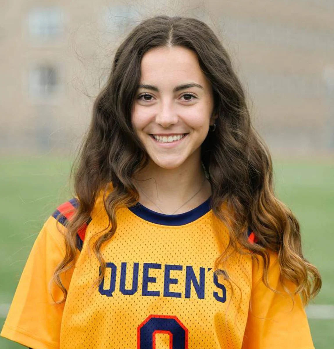 Young woman with long, wavy brown hair smiling outdoors, wearing a yellow sports jersey with blue and red accents that reads 'Queens' and the number zero.