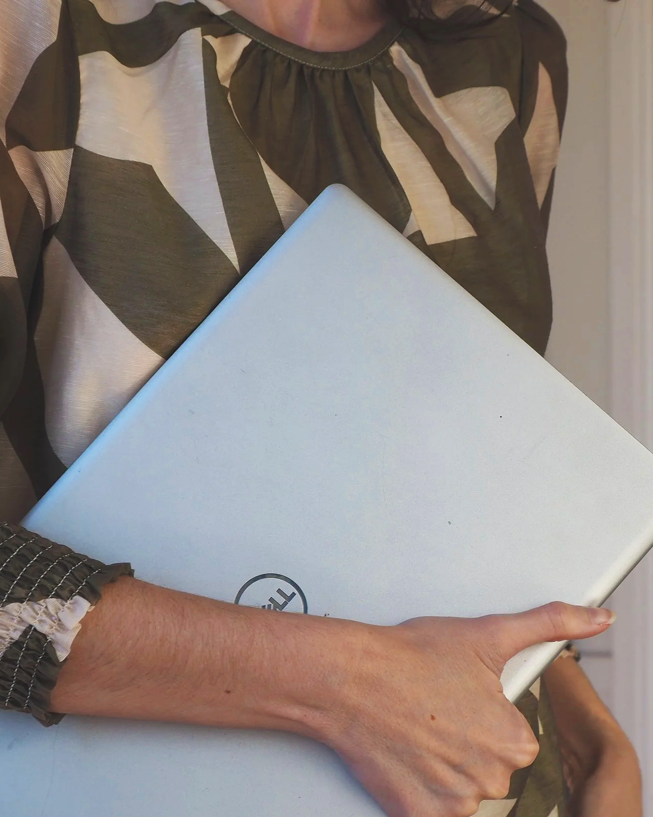 Person holding a silver Dell laptop, wearing a patterned long-sleeve shirt, arm extended over a surface.