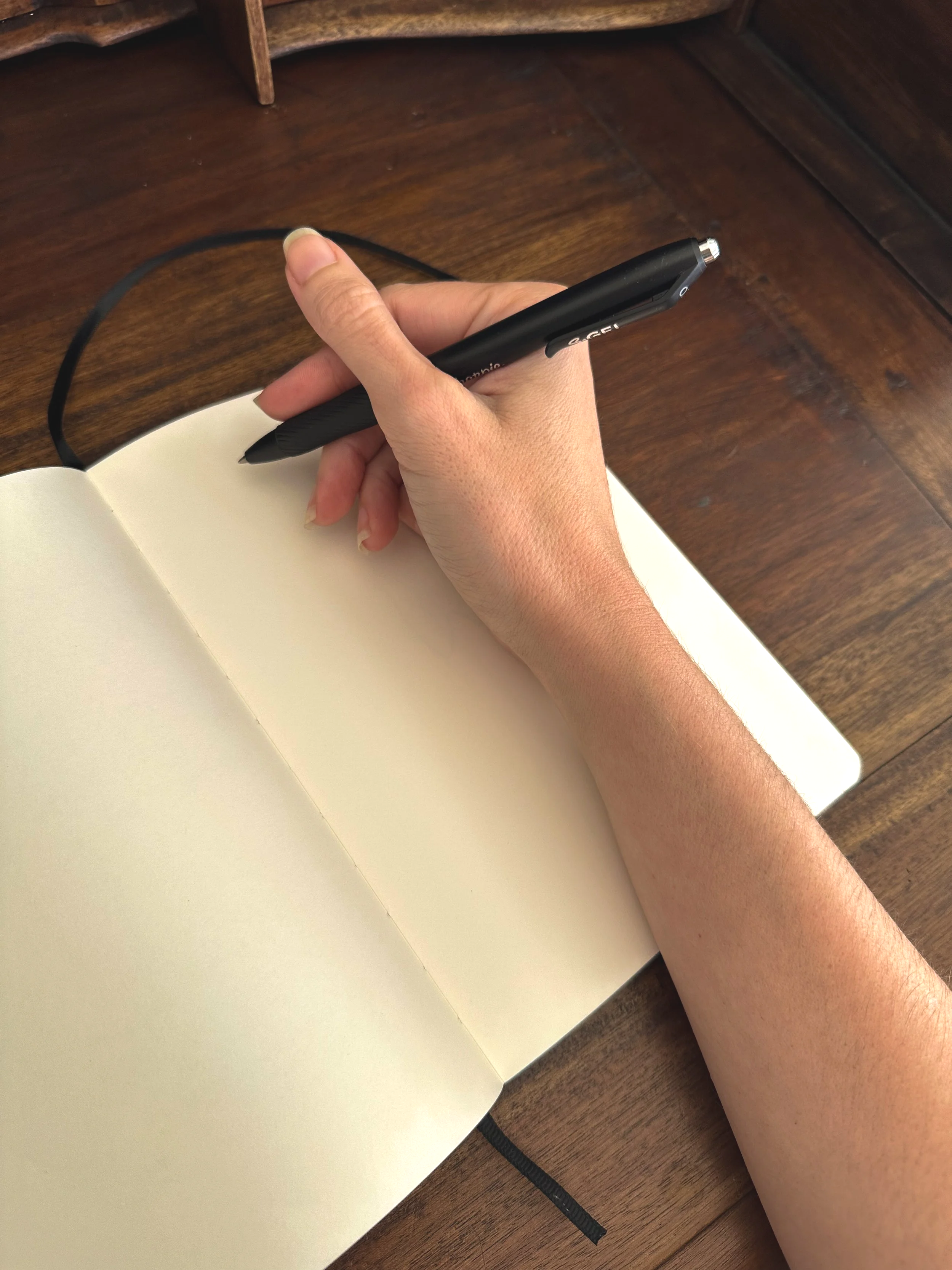A person holding a black pen over an open blank notebook on a wooden desk.