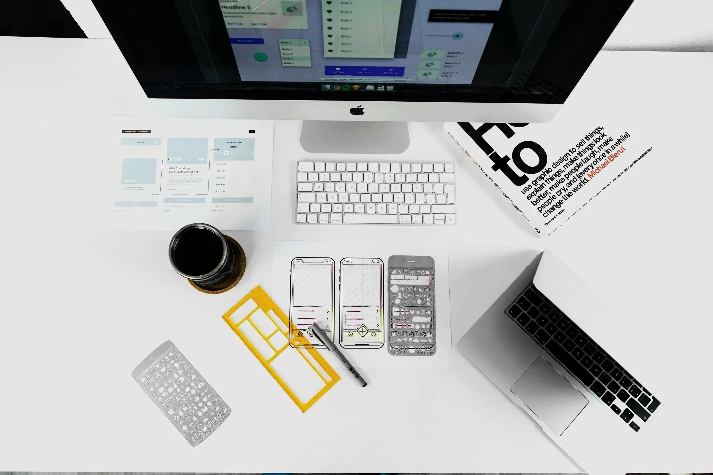 Overhead view of a white desk with a computer monitor, keyboard, pen, paper diagrams, and small electronic components. A newspaper with a quote is partially visible on the right side.