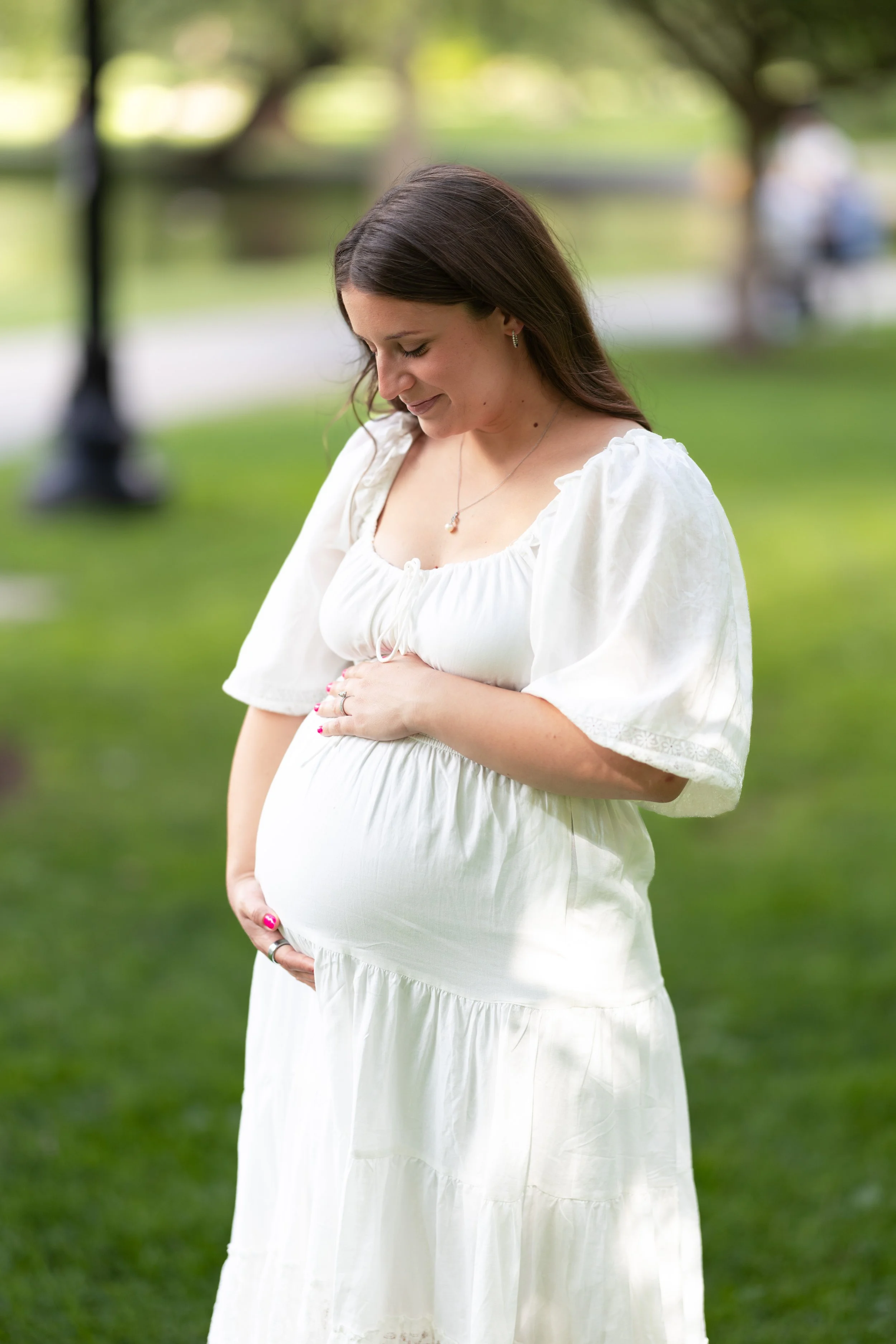 Portrait of a pregnant woman in a white dress outdoors, cradling her belly with one hand, smiling and looking down thoughtfully.