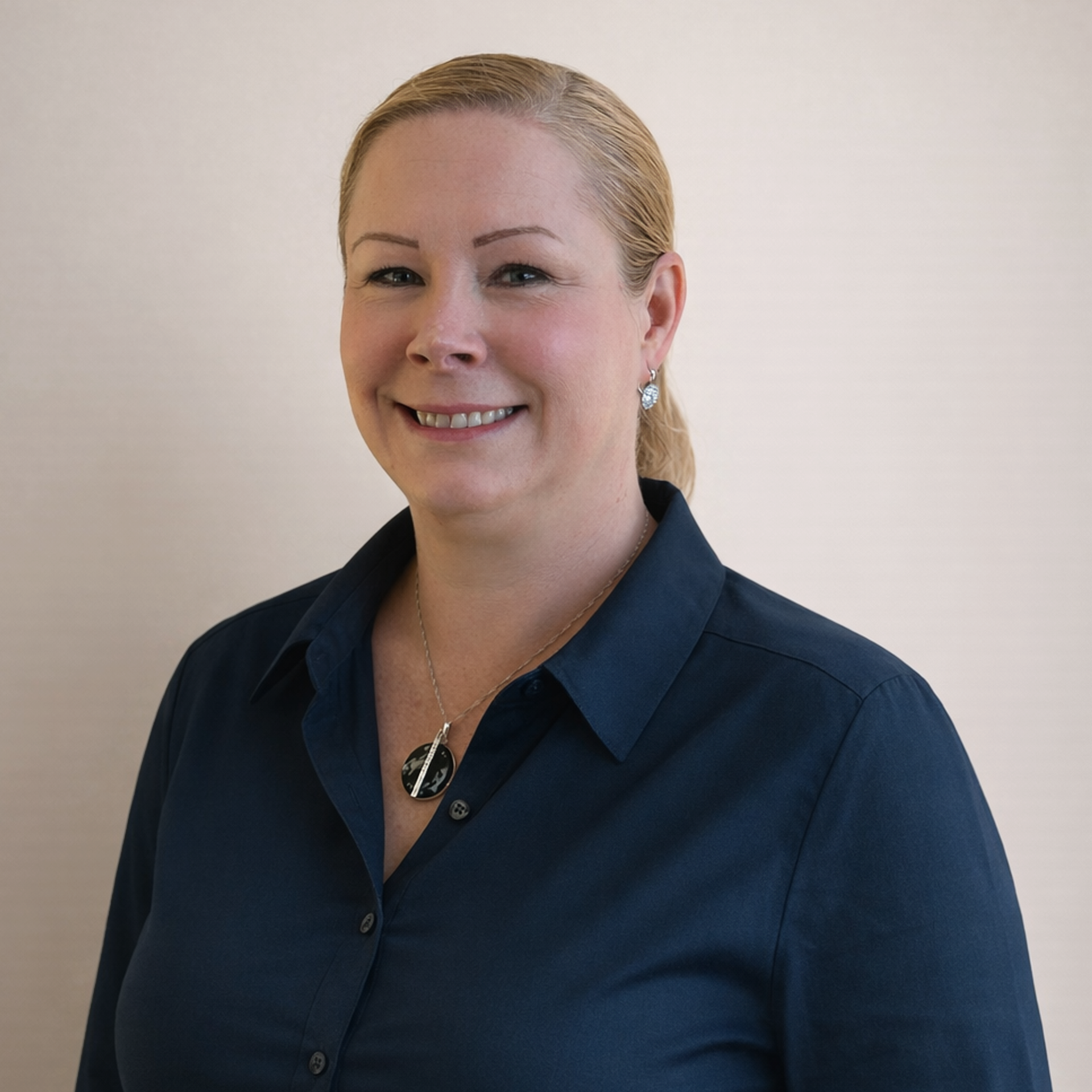 Close-up of a woman with blonde hair slicked back, wearing a dark blue collared shirt, silver earrings, and a necklace with a black and silver pendant, smiling against a plain light background.