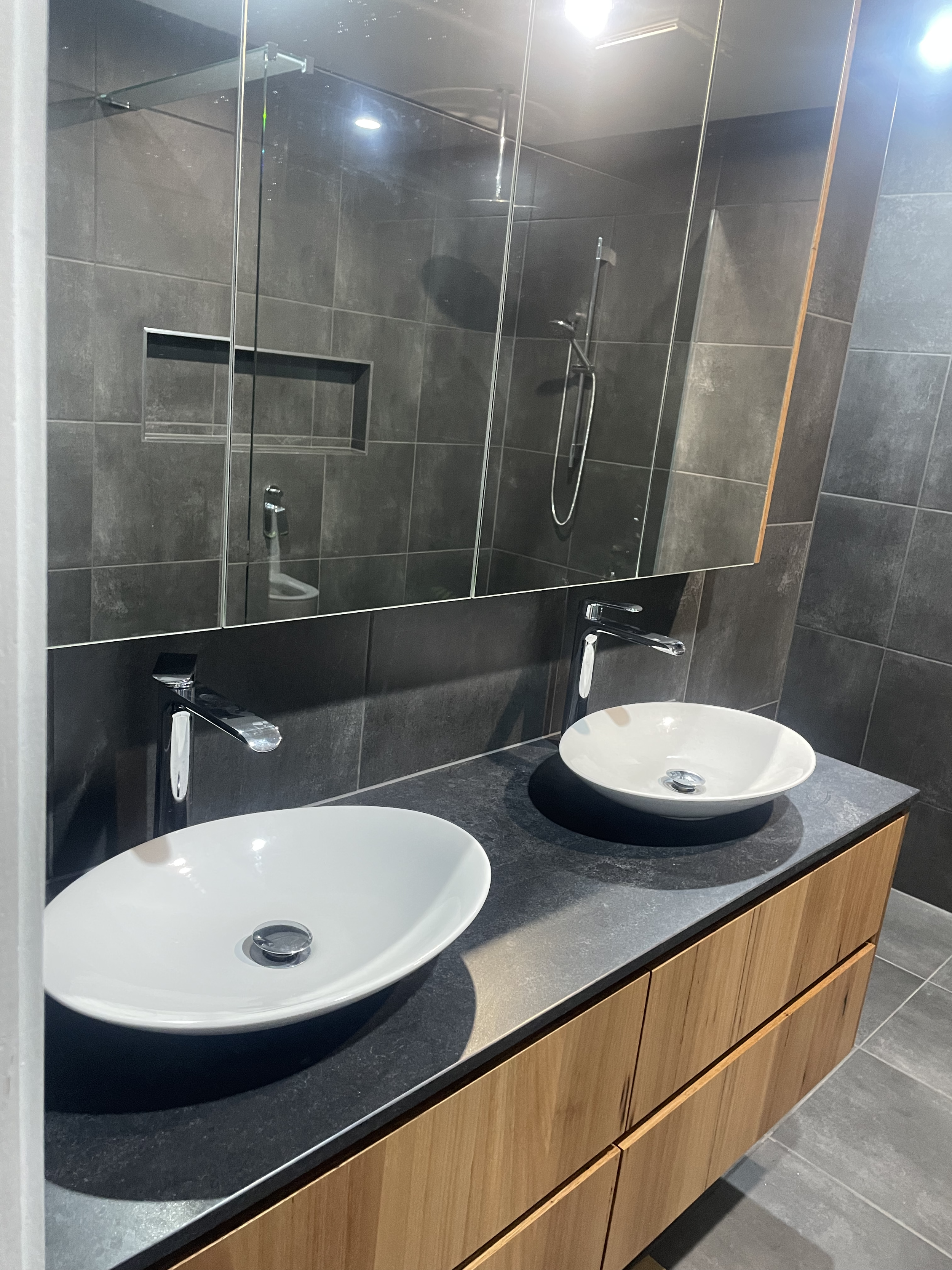 A modern bathroom with two white oval sinks on a dark gray countertop, wooden cabinets below, a large mirror above, and a glass-enclosed shower with a handheld showerhead and gray tiled walls.