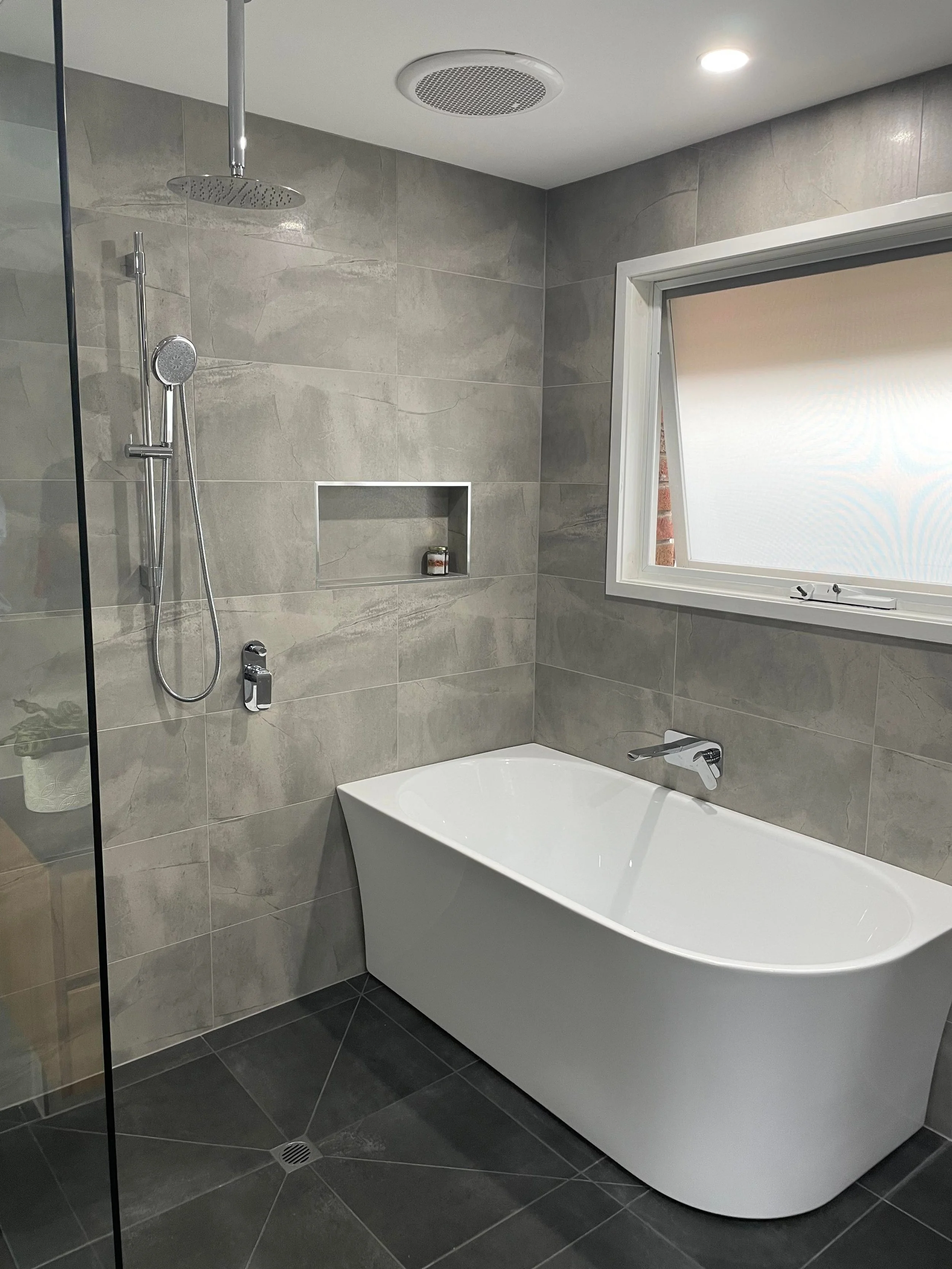 Modern bathroom with gray tile walls, a white bathtub, a large window, and a shower area with a rainfall showerhead and handheld shower attached to the wall.