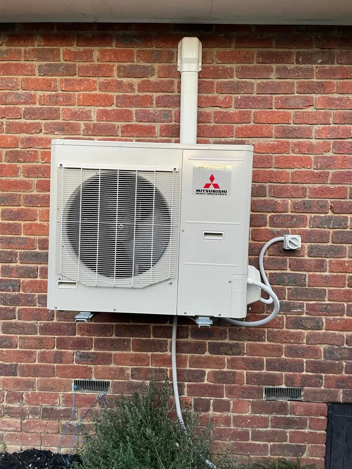Outdoor air conditioning unit mounted on a brick wall, with electrical wiring and conduit attached.