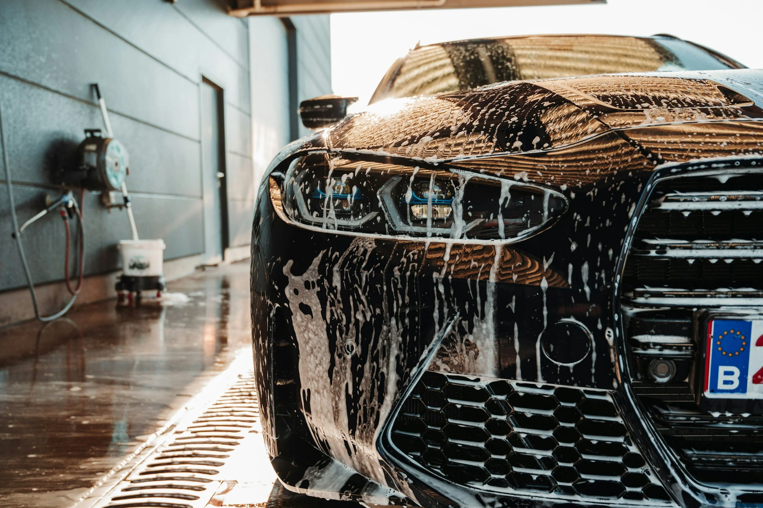 A black car being washed with soap and water at an outdoor car wash station, sunlight reflecting off the wet surface.