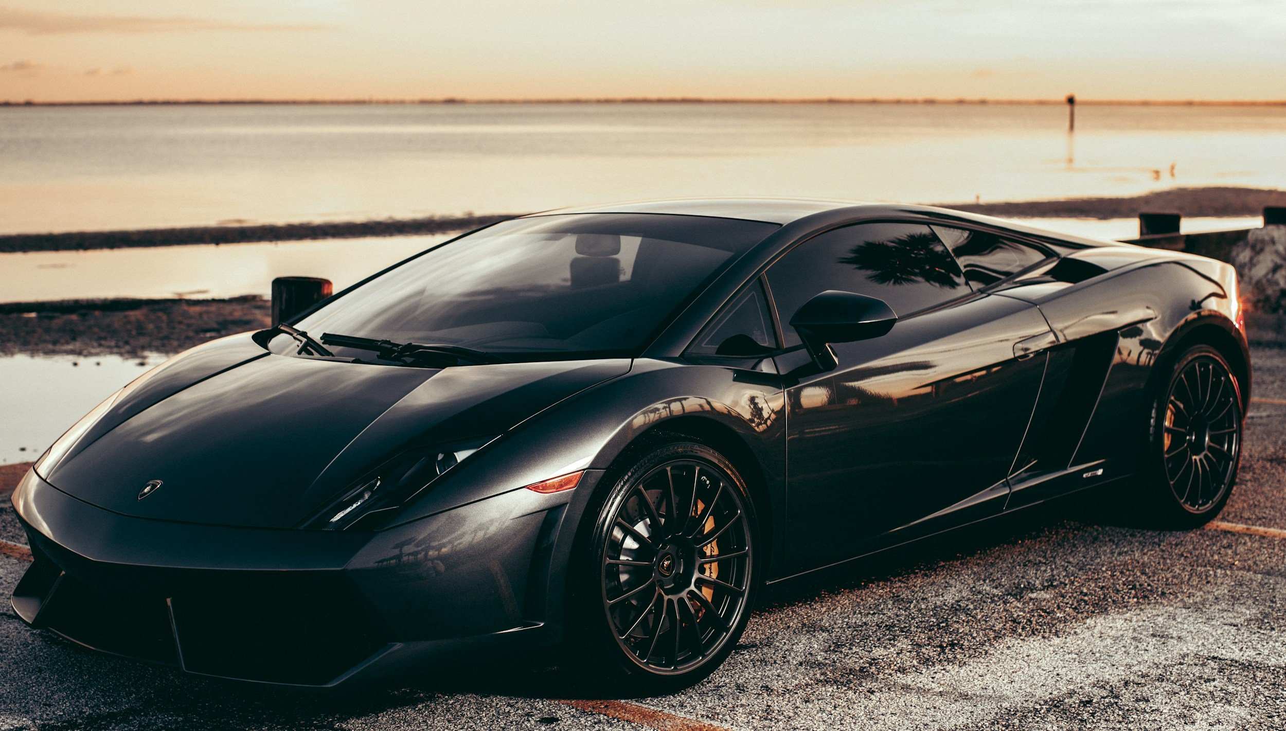 A sleek black sports car parked near a body of water at sunset, with reflections of trees and the sky on its shiny surface.