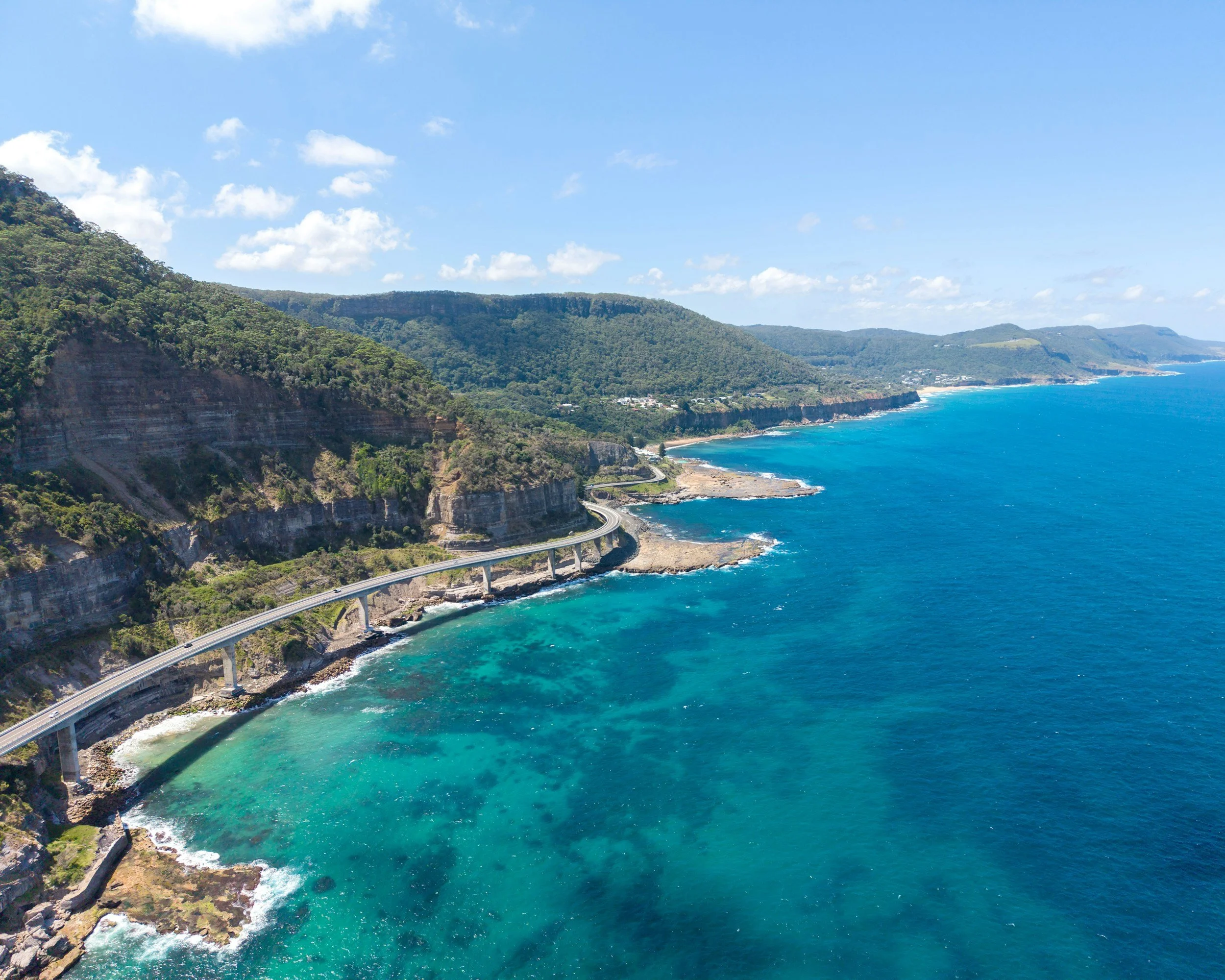 Coastal scenery with cliffs covered in lush green trees, a winding road along the cliffs, and the ocean with blue water and white waves under a partly cloudy sky.
