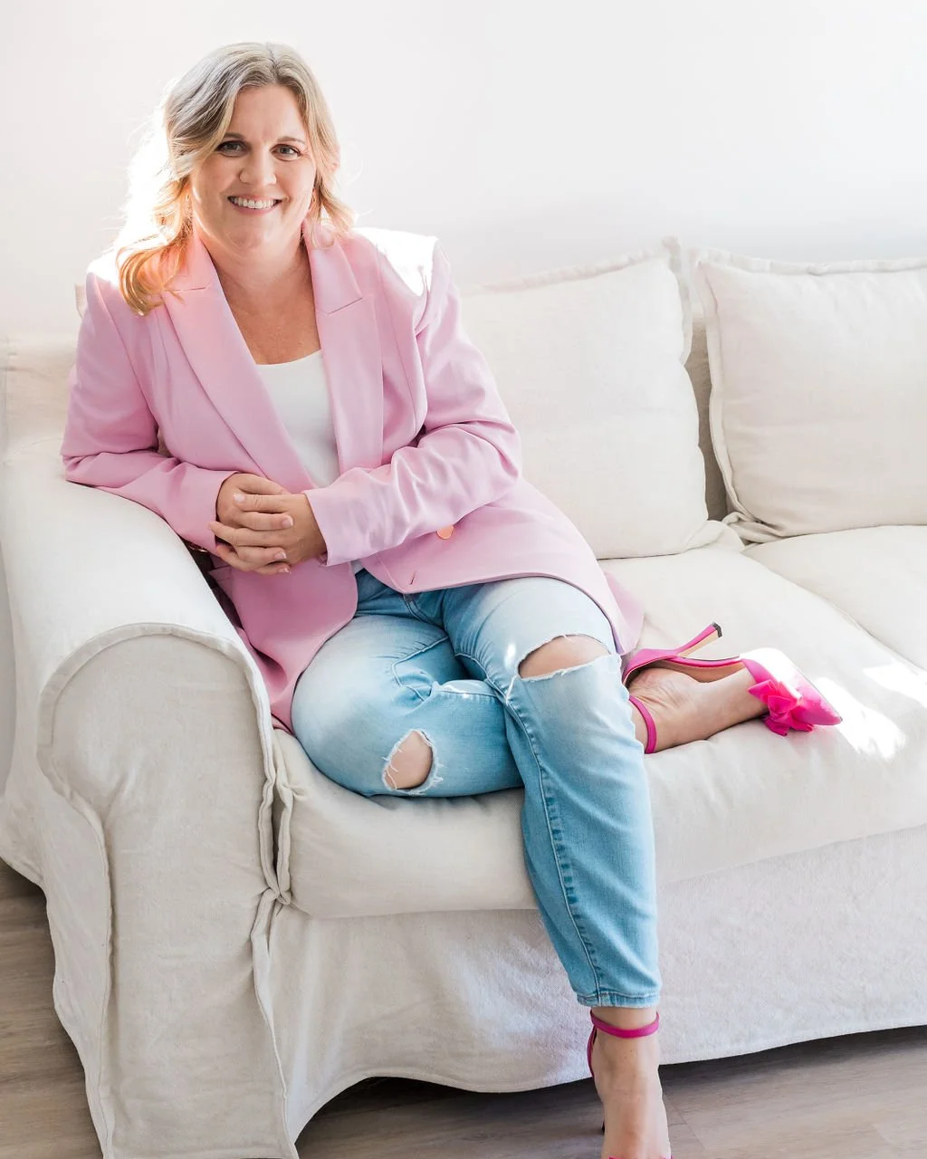 A woman with shoulder-length blonde hair sitting on a white sofa, wearing a light pink blazer, ripped jeans, and bright pink high heels with bows, smiling at the camera.