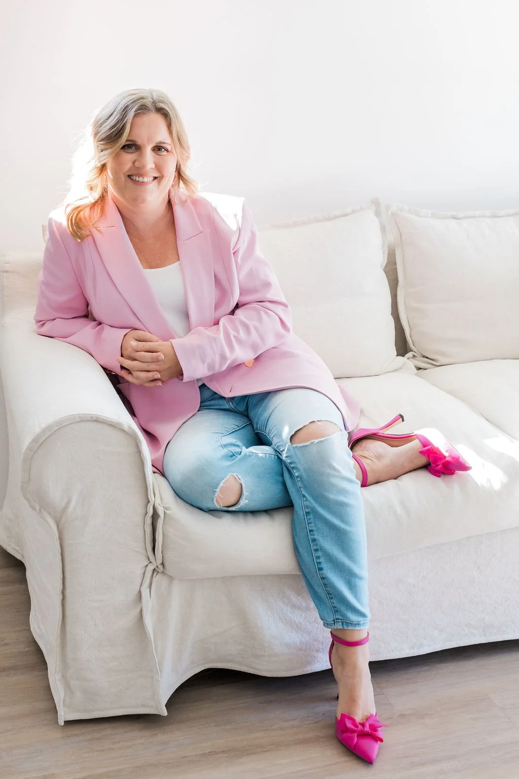 A woman sitting on a cream-colored couch, wearing a pink blazer, distressed jeans, and pink high heels with bows, smiling at the camera.