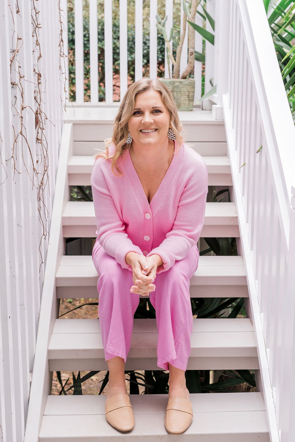 A woman with blonde hair wearing a pink cardigan and matching pants, sitting on outdoor white stairs, smiling at the camera, surrounded by white railings and greenery.