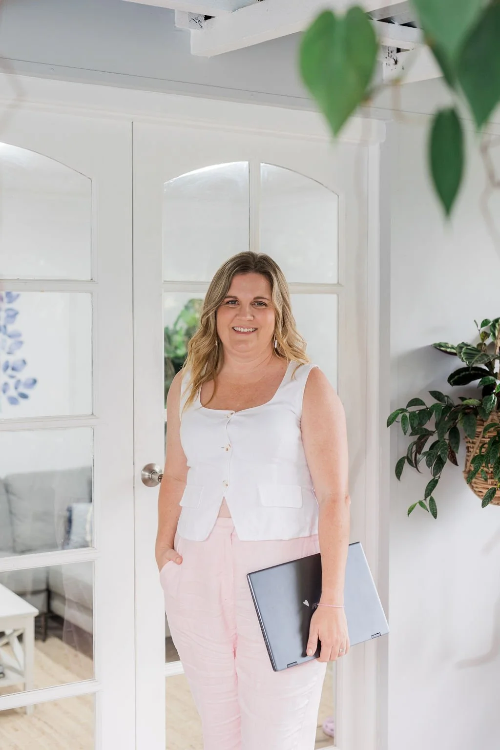A woman standing indoors holding a closed laptop, smiling, with a white door and green plants in the background.
