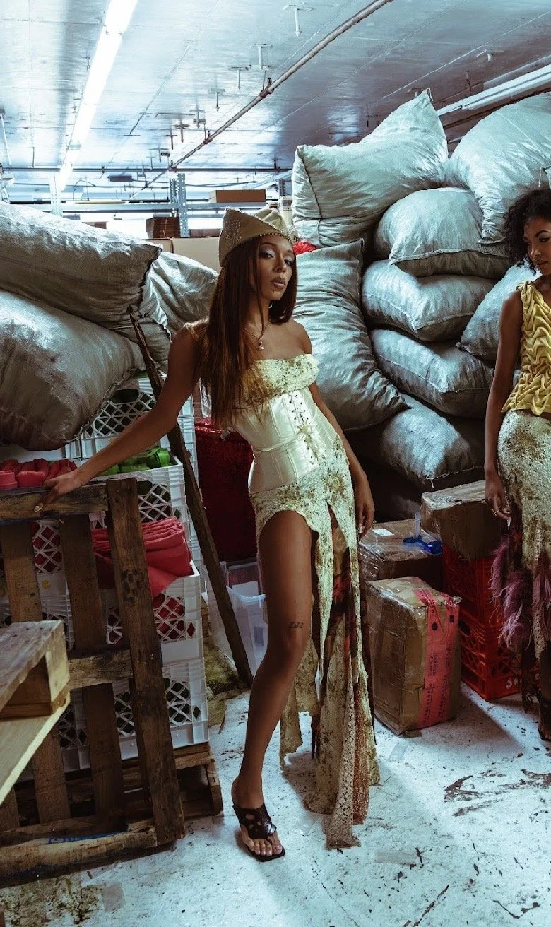 A woman dressed in a long, vintage-inspired dress with a slit, standing in a warehouse surrounded by large bags and boxes.