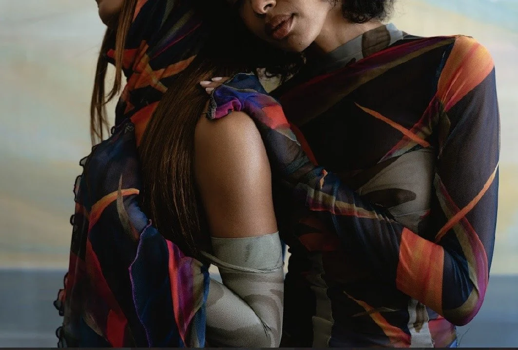 Two women embrace, one holding the other's leg, both wearing colorful, patterned long-sleeve tops.