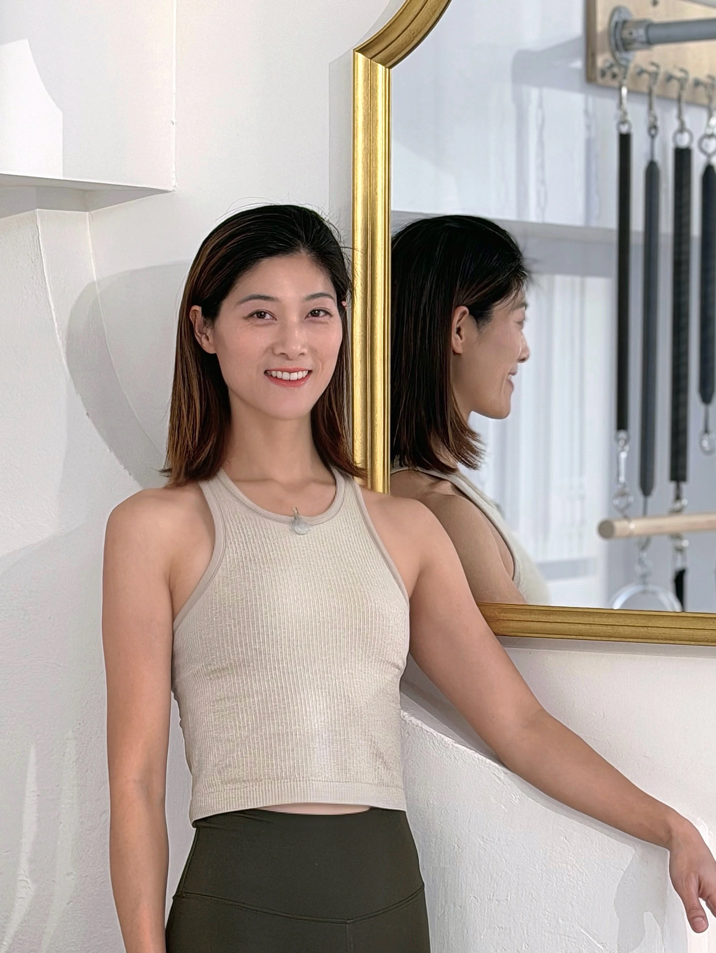 A woman with shoulder-length dark hair wearing a beige tank top and black pants standing in front of a mirror with a gold frame. The mirror reflects her profile and a wall with hanging exercise equipment.