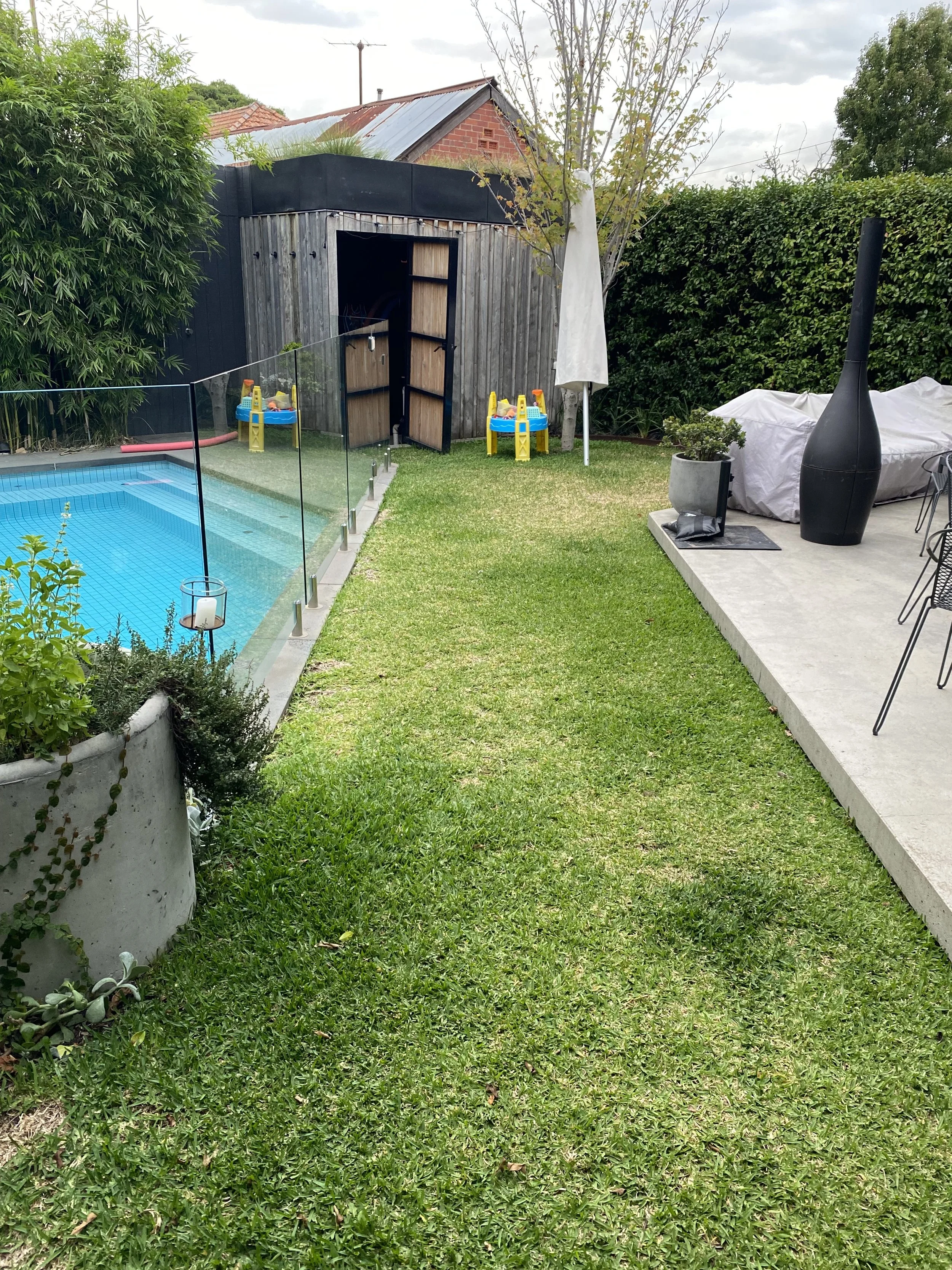 A backyard scene with a grassy lawn, a swimming pool with glass fencing, a small play area with yellow and blue toys, a white patio umbrella, potted plants, a covered object, and a black outdoor heater, surrounded by greenery and a red brick house in the background.